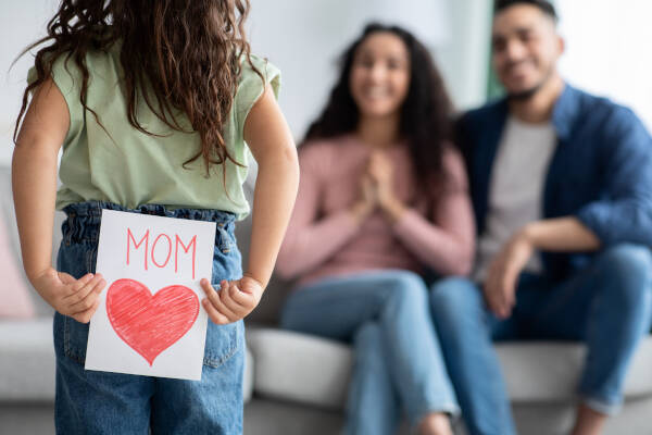 Girl hides MOM card behind her back, mom sits on sofa with her dad in anticipation