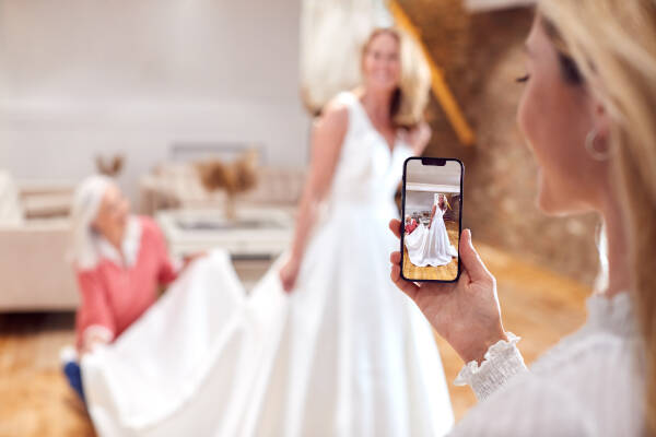 Grandmother, adult daughter, and granddaughter try on wedding dress, taking picture on focused phone