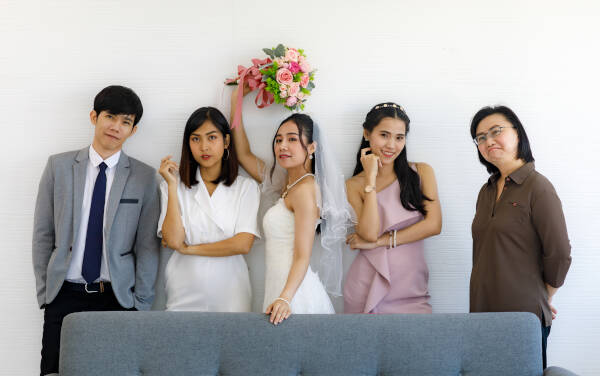 Groom in suit, bride in wedding dress with veil, holding pink roses, smile behind gray sofa with bridesmaid and auntie