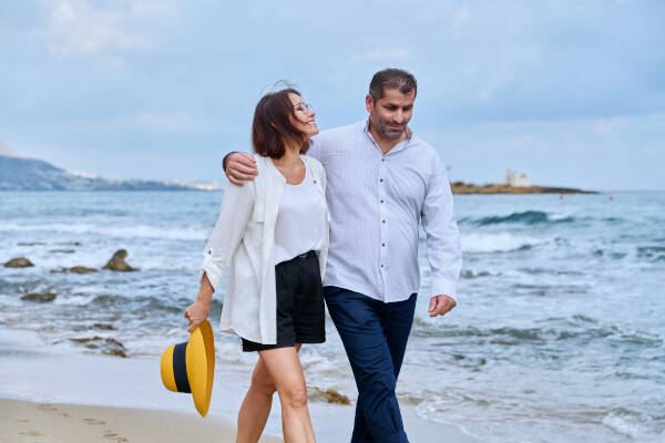 Happy mature couple walks on beach with sea in background. Wife wears glasses and holds yellow hat.