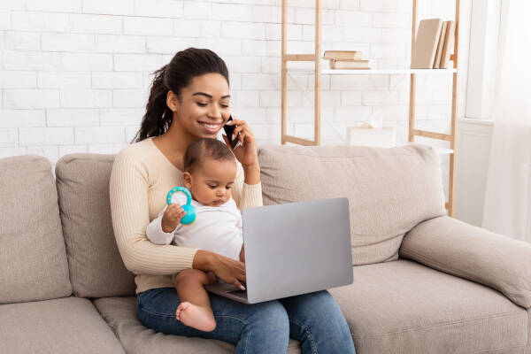 Happy mother with baby in lap chats on phone, multitasking with laptop on couch at home