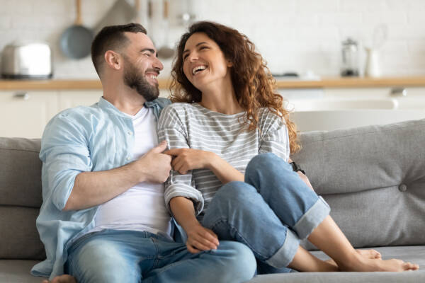 Happy young couple hugging, sitting on a cozy couch together, overjoyed laughing woman and man having fun