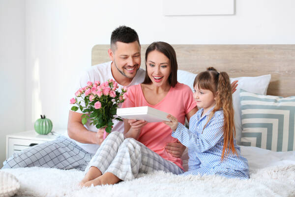 Husband and little daughter surprise woman with a Mother's Day gift while she sits on the bed