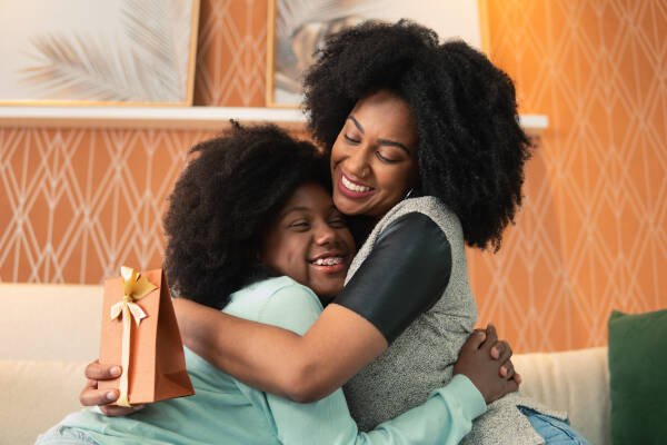 In the living room, a mom beams with appreciation as her teenage daughter hands her a Mother's Day gift