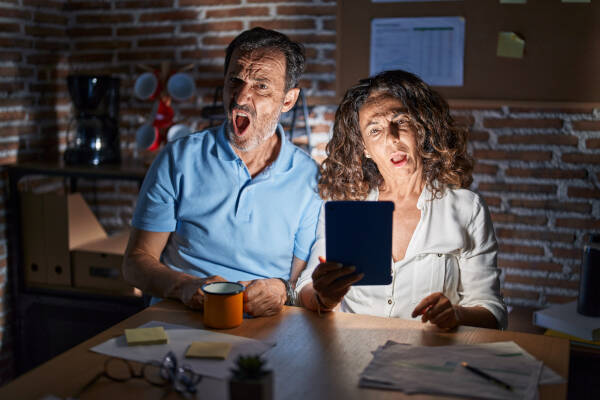 Middle-aged couple, shocked faces illuminated by touchpad screen, sitting at table at night