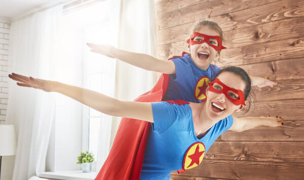 Mother and child play together, with the child pretending to fly on mom's back. Both wear superhero costumes