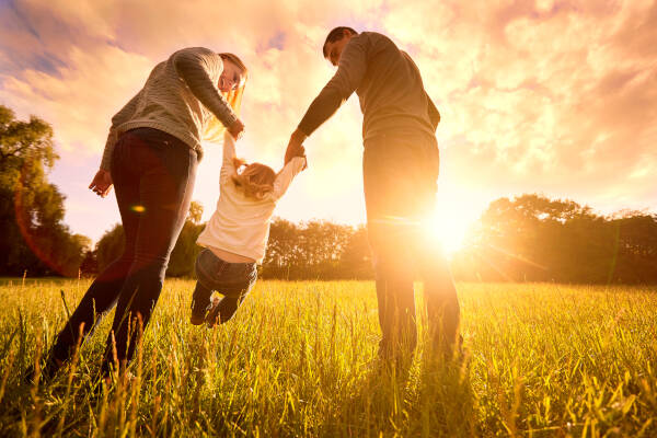 Mother and father swing their young child between them in a field at sunset