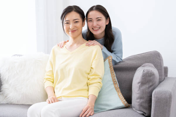 Mother sits on the sofa as her daughter hugs her from behind, both smiling at the camera