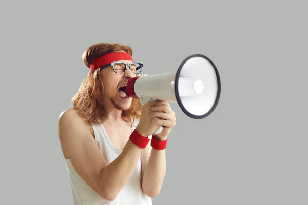 Nerdy young man in retro sportswear stands isolated on gray background, yelling into megaphone with enthusiasm
