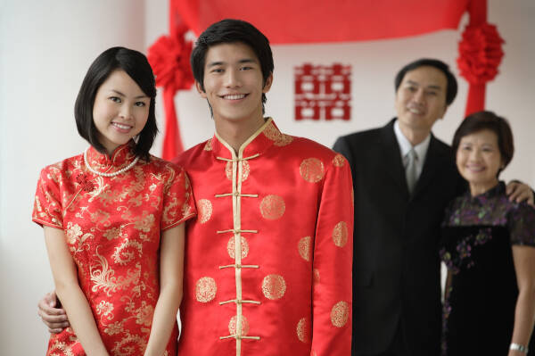 Newlywed couple smiles at the camera while aunt and uncle watch from a distance.