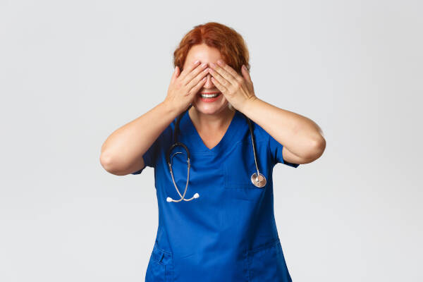 Nurse with stethoscope around neck covers her eyes playfully, smiling warmly.