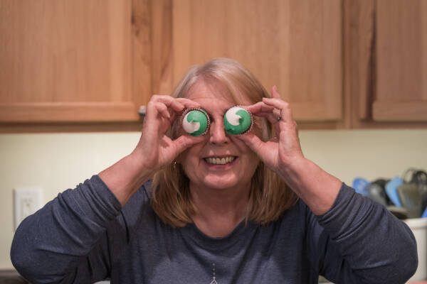 Older woman holds two mini frosted cupcakes up to her eyes, acting silly.