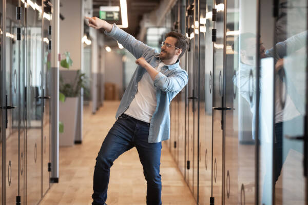 Overjoyed male employee in glasses performs a victorious dance in a modern office hallway