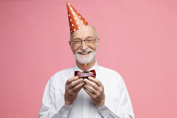 Senior man celebrates 80th birthday, holding cake with candle, smiling against pink backdrop