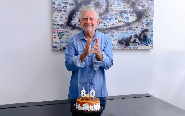 Senior man celebrating his 80th birthday, standing behind a cake with 80 number candles on a dining table.