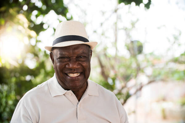 Senior man, wearing a white hat, stands outdoors, smiling warmly at the camera, radiating happiness