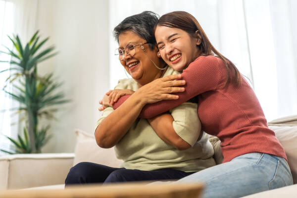 Senior mother and adult daughter share a warm hug on the sofa at home, both wearing bright smiles.