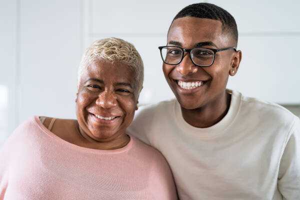 Senior mother and her young adult son pose together for a photo inside their home, both wearing smiles