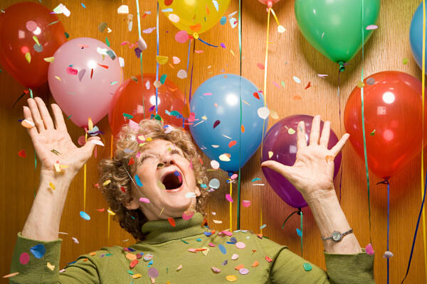 Senior woman cheers at a party, surrounded by balloons and falling confetti, embracing the joyous atmosphere