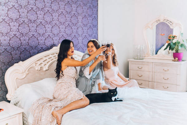 Sisters sit on their bed, clinking glasses in celebration of one of their engagements