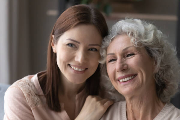 Smiling adult daughter-in-law hugs the shoulders of her older mother-in-law, they look aside and dream together