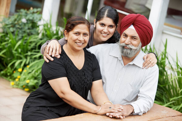 Smiling parents sit on bench outdoors as daughter wraps her arms around them from behind