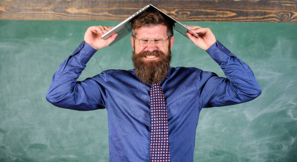 Teacher grits teeth in frustration, holding laptop as a roof over head, with chalkboard background