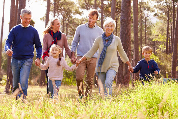 Three generations of family walk through the woods, smiling, with trees in the background and grass in view.