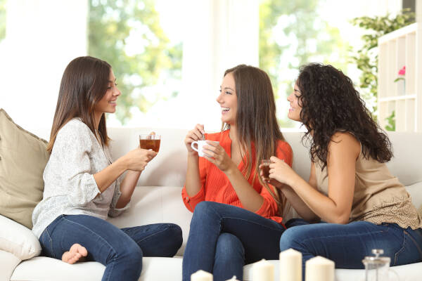 Three happy friends chatting and enjoying coffee and tea while sitting on a couch at home