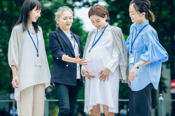 Three women engage in conversation with a pregnant colleague outdoors at work