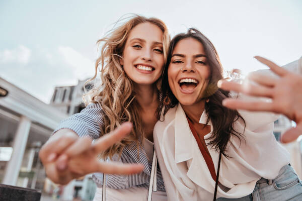 Two girls making fun gestures to the camera, posing outdoors against a sky background.