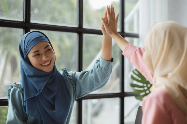 Two sisters high-five each other indoors in front of a window, both wearing smiles.