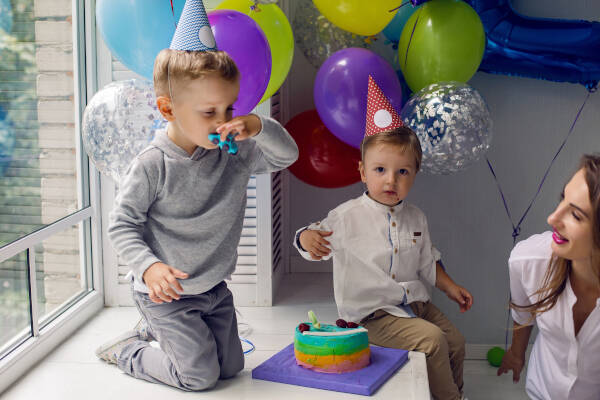 Two-year-old boy kneels on a window sill with his brother and mother, birthday cake on the ledge, and balloons on the sides.