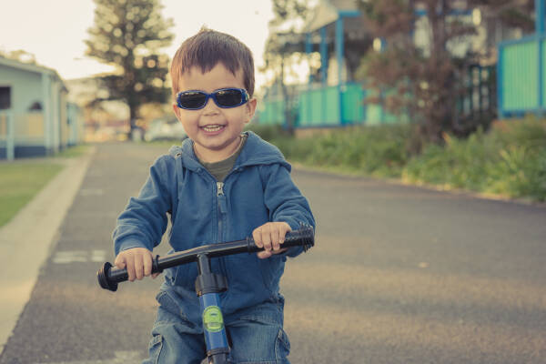 Two-year-old boy rides his bike in a caravan park, wearing sunglasses and smiling.