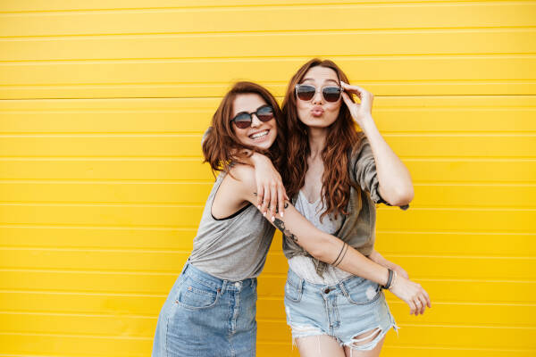 Two young, happy women friends standing over a yellow wall, looking at the camera and blowing kisses.