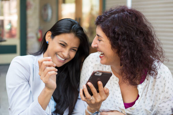 Two young women share a laugh while looking at a smartphone together.