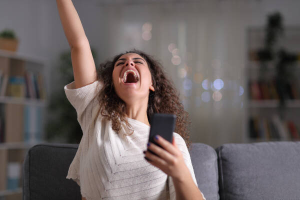 Woman celebrates success, holding mobile phone, sitting on couch at night at home, expressing excitement and joy