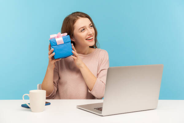 Woman employee at desk holds gift near ear, laptop and mug nearby, eagerly listening for its contents