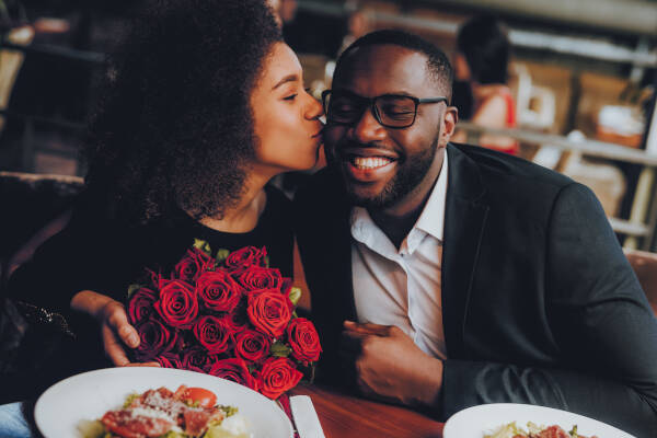 Woman kisses her boyfriend on the cheek while holding roses in a restaurant, man is beaming