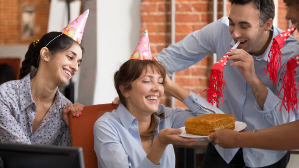 Work team in party hats presents a surprise cake to their boss; a couple of employees blow party horns