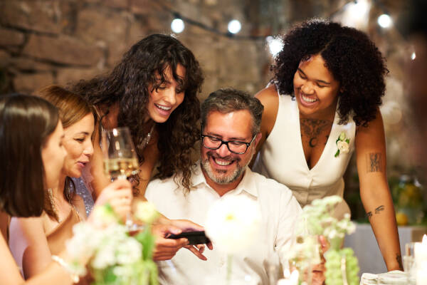 Young brunette bride happily showing photos on her smartphone to her uncle, with family gathered around the table