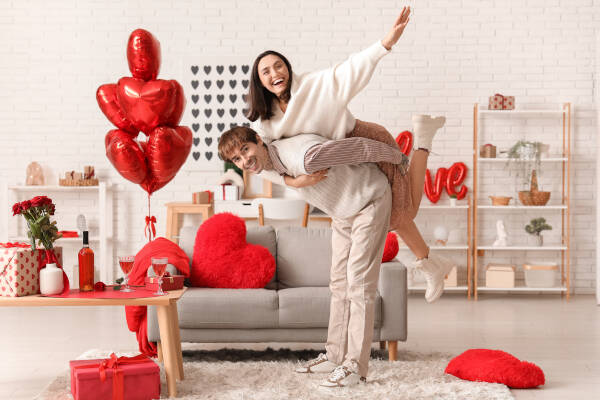 Young couple smiles, wife on husband's back, heart-shaped balloons and 'love' balloon in background