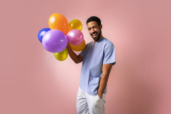 Young dentist happily holds balloons over a pink background, radiating joy and positivity