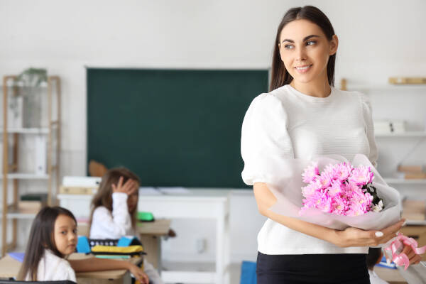 Young female teacher holds bouquet of flowers in classroom, children visible in the background