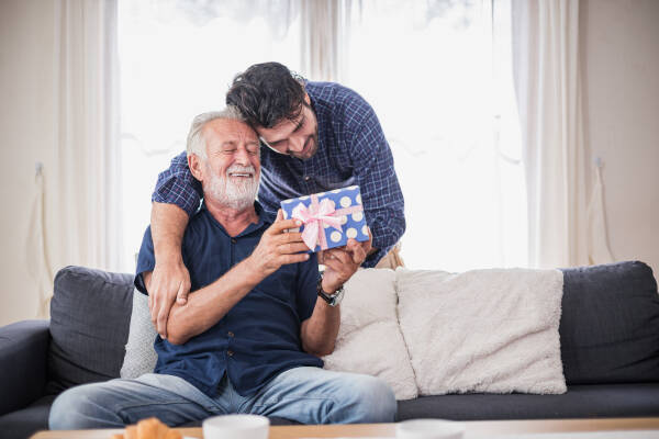 Young man bends down to hug his dad, who is sitting on the sofa, and presents him with a spotty-wrapped gift
