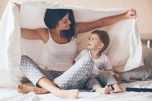 Young mother and her two-year-old son, both dressed in pajamas, are relaxing and playing under bed sheets.