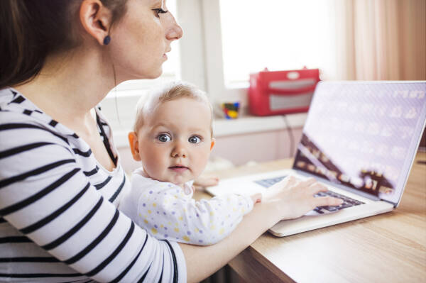 Young mother multitasks in home office, working on computer with her baby daughter in her lap