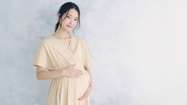 Young pregnant woman stands, cradling her belly with a smile against a serene off-white backdrop