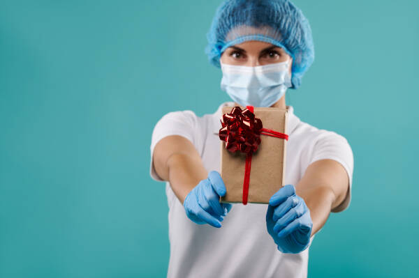 Young woman dentist, wearing hat and mask, holds a brown gift with red bow up to camera against a turquoise backdrop.