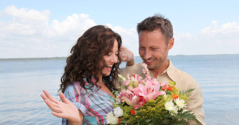 Man surprises woman with a bunch of flowers on the beach, with the sea in the background.